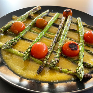 Espárragos verdes a la plancha con tomates cherry asados, servidos sobre una base de salsa de níspero, presentados en plato negro.