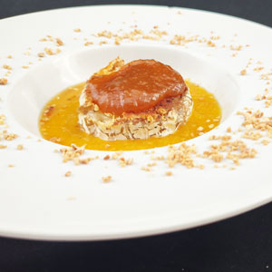 Queso tibio de cabra con costra, servido sobre salsa de níspero y decorado con chispas de cacahuete crocante en plato hondo blanco.