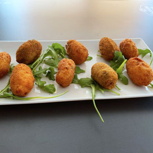 Bandeja rectangular con un surtido de croquetas variadas sobre un lecho de hojas de rúcula, servida sobre una mesa oscura.