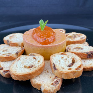 Paté de oca coronado con mermelada de níspero y decorado con hoja de menta, acompañado de tostadas de pan con pasas, servido en plato negro.