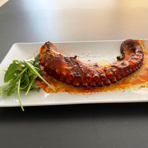 Pata de pulpo a la brasa servida sobre plato rectangular blanco, acompañada de brotes verdes y aliñada con sal, aceite y pimentón.