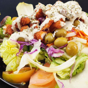 Ensalada tipo César con lechuga, col lombarda, tomate, aceitunas verdes, crujientes de pollo empanado y salsa césar, servida en plato negro.