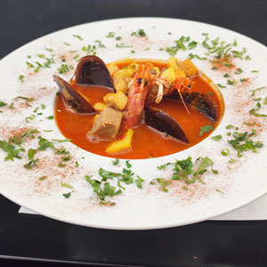 Sopa de mariscos con mejillones, gamba, dados de pescado y picatostes, servida en plato hondo blanco y decorada con perejil y pimentón.