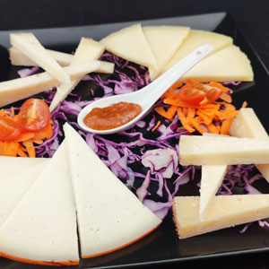 Tabla de quesos valencianos variados servida sobre col lombarda rallada, zanahoria, tomate cherry y mermelada de níspero en cuchara de cerámica, en plato negro.