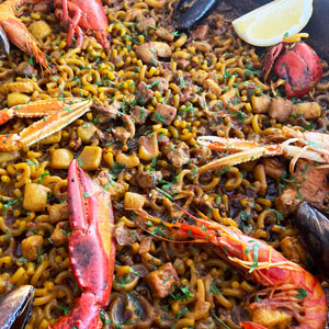 Fideuà de marisco con bogavante, cigalas, gambas, calamar y mejillones, cocinada en paella y decorada con perejil y limón.